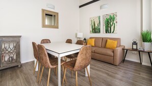 Een eetgedeelte met een witte tafel en bruine stoelen, een bruine bank met gele kussens, en twee schilderijen van cactussen aan de muur.