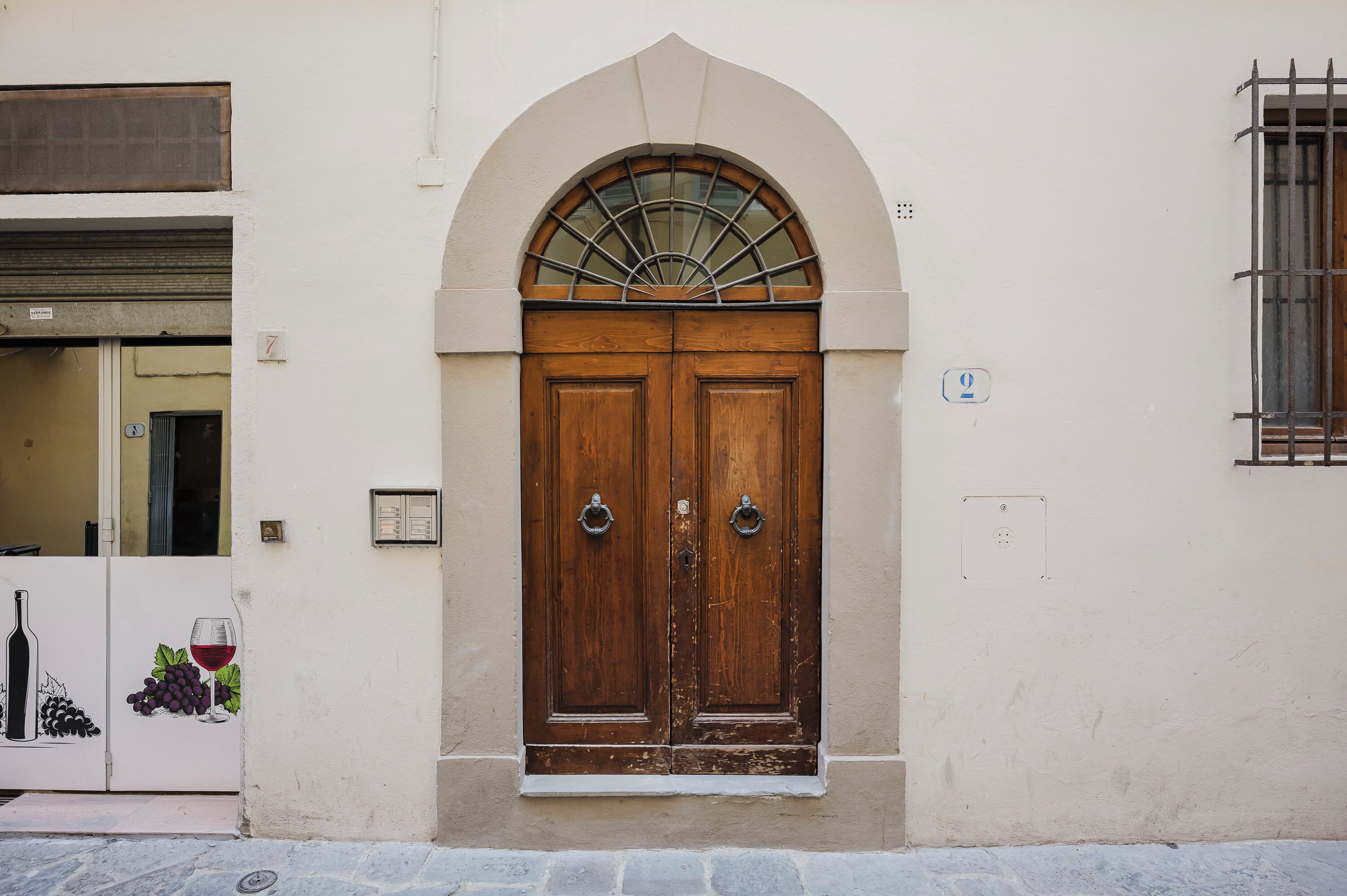 A double wooden door with a semi-circular window above it, set in a plain wall with a small window to the right.
