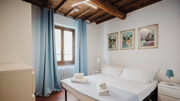 Una habitación de hotel con cama, mesita de noche, lámpara, ventana con cortinas y dos cuadros en la pared.