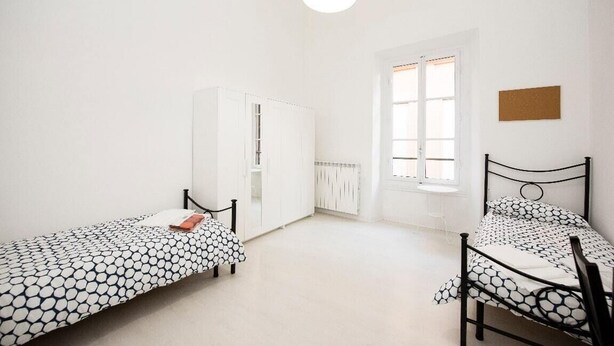 A bedroom with two beds, a wardrobe, a window, and a hanging light fixture.