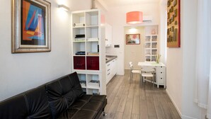 Uma sala de estar moderna com um sofá de couro preto, uma estante branca com livros e objetos decorativos, e um espaço de jantar com mesa e cadeiras.