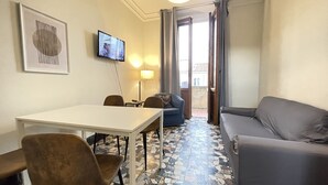 A room with a white table, two chairs, a grey sofa, a blue sofa, a TV on the wall, and a patterned floor. - Fortezza Fourth Floor 4 Pax (Firenze)
