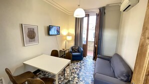 A compact room with a white table, two chairs, a blue armchair, a grey sofa, a TV on the wall, and a patterned floor. - Fortezza Fourth Floor 4 Pax (Firenze)