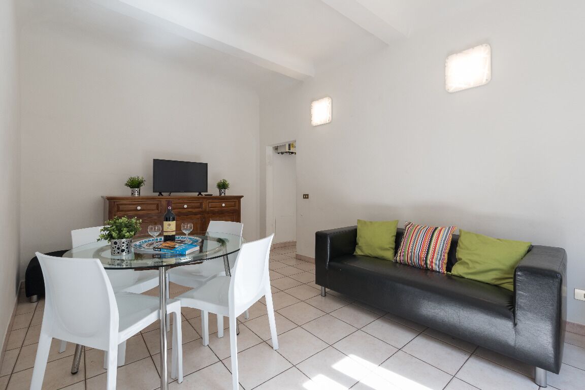 Ein modernes Wohnzimmer mit einem Glastisch, weißen Stühlen, einem schwarzen Sofa mit bunten Kissen, einem Fernseher auf einer Holzkommode und einer kleinen Pflanze.