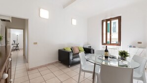 Un moderno espacio de estar con una mesa de comedor de vidrio, sillas blancas, un sofá de cuero negro con cojines de colores, una mesita auxiliar y una ventana con persianas.