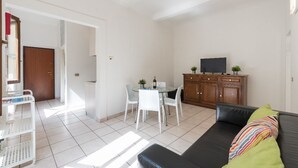 Una sala de estar con un sofá de cuero negro, una mesa de comedor blanca con sillas, un mueble de madera con un televisor y una puerta que da a otra habitación.