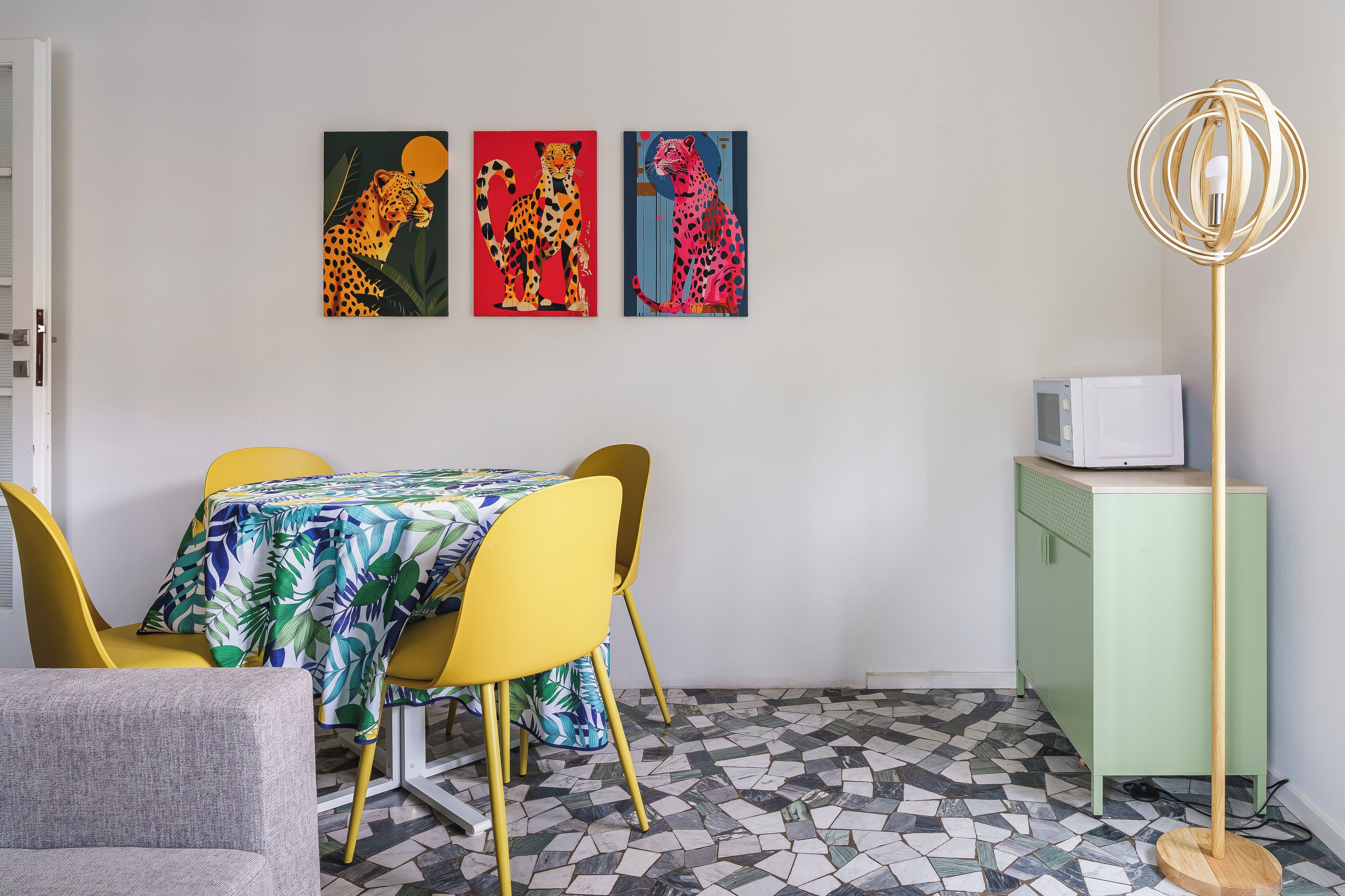 A dining area with a table covered in a tropical leaf tablecloth, yellow chairs, and a green cabinet with a white appliance on top.