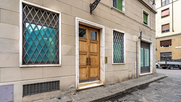 A building with a wooden double door, a window with a metal grid, and a small vent on the wall. - Buonarroti Apartment (Firenze)