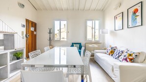 A modern living room with a dining table, white chairs, a sofa with colorful cushions, and a blue accent chair. There are two windows providing natural light, and a door with a coat rack. - Faenza 73 - Three Bedroom Apartment, Sleeps 5 (Firenze)