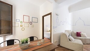 A cozy living room with a wooden dining table, a sofa, and a wall with geometric shapes and a city skyline mural. - Faenza double - One Bedroom Apartment, Sleeps 2 (Firenze)