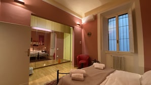 A bedroom with a bed, a red chair, a window, and a mirror. - Battistero Apartment (Firenze)