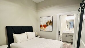 Un dormitorio con una cama grande, un ventilador de techo, una mesita de noche, un cuadro colgado en la pared y un baño visible a través de una puerta abierta.