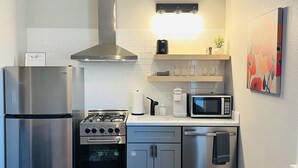Cocina compacta con refrigerador, estufa, horno, microondas y lavavajillas de acero inoxidable. Hay una repisa de madera con frascos y una planta pequeña. Un cuadro está en la pared, y un sofá negro se encuentra en primer plano.