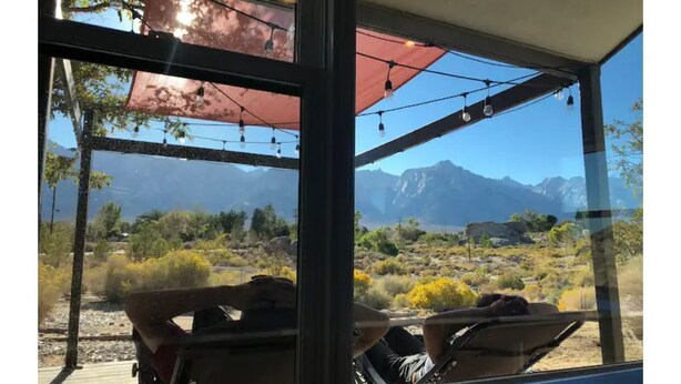 A room with a large window offering a view of a desert landscape and mountains. There are two chairs with cushions and a table. - >>MAGICAL property on 5 acres in the Alabama Hills (Lone Pine)