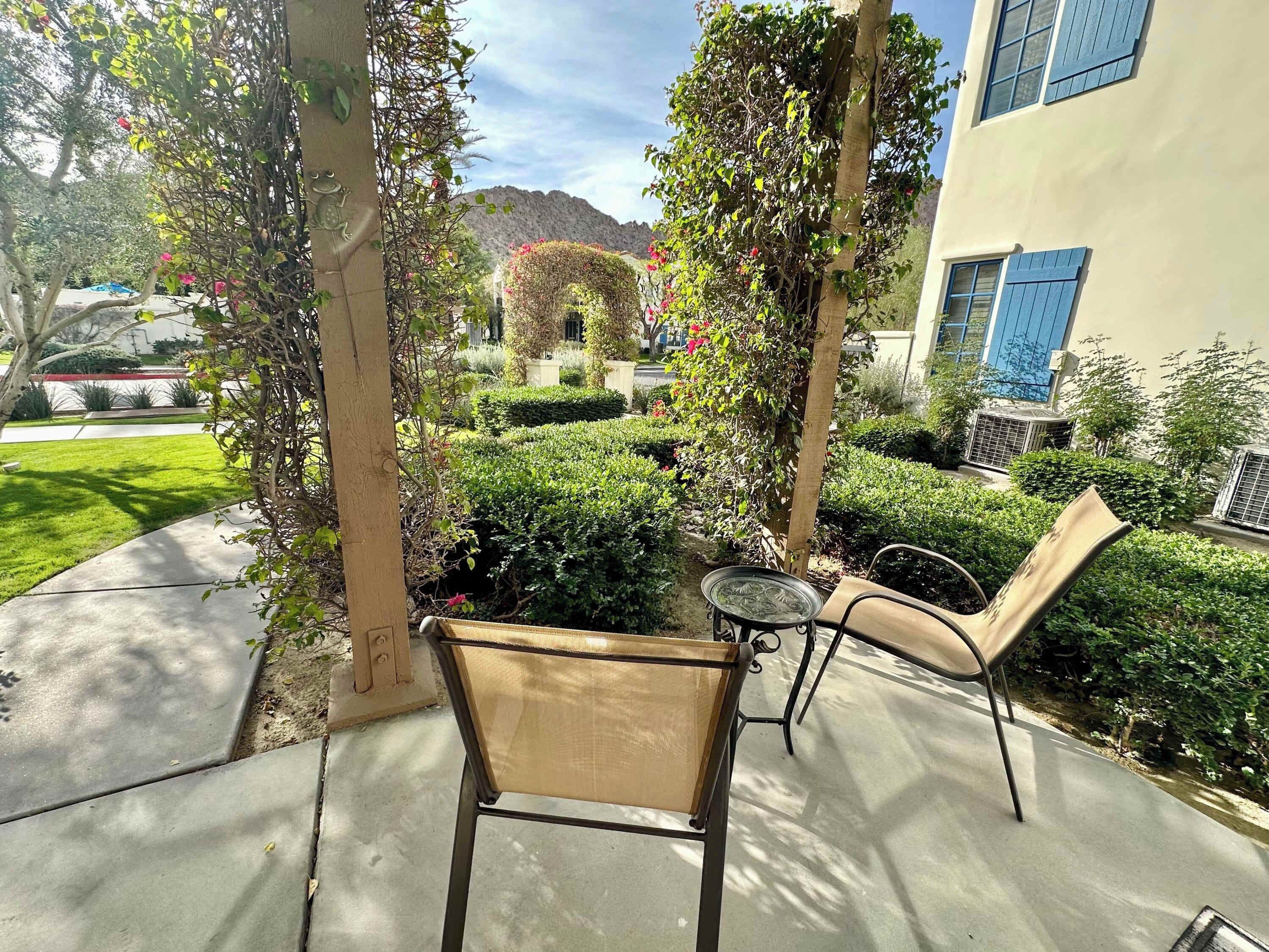 A covered patio area with a chair and table, surrounded by greenery and a building with blue shutters.