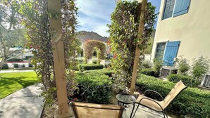 Un patio couvert avec une chaise et une table, entouré de verdure et bordé d’un bâtiment aux volets bleus.