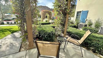 A covered patio area with a chair and table, surrounded by greenery and a building with blue shutters.
