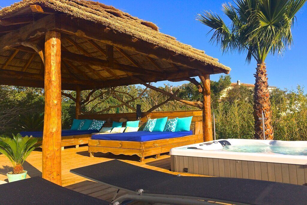 A wooden outdoor seating area with a thatched roof, a hot tub, and a bed with blue cushions.