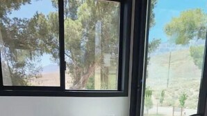 A room with a bed, a wooden bench, a small table with a glass, and a view of trees and a landscape through large windows.