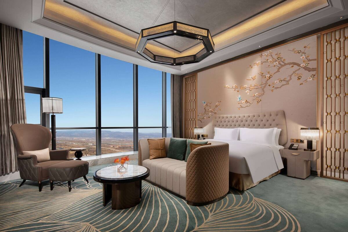 A hotel room with a large window, a bed, a sofa, a chair, a side table, and a decorative wall piece.