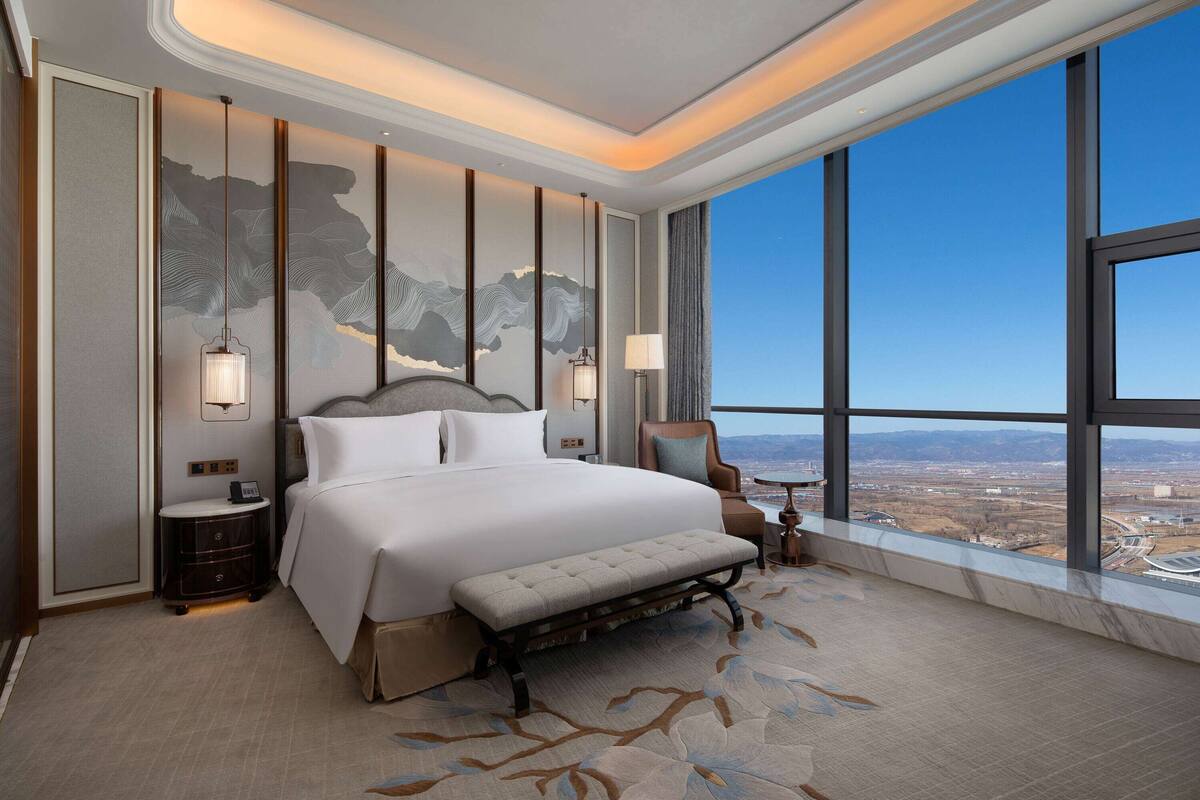 A hotel room with a large bed, a bench, a nightstand, a chair, and a view of the city through large windows.