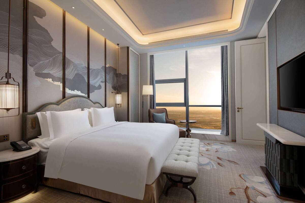 A hotel room with a large bed, a television, a desk with a lamp, and a view of the desert through a large window.
