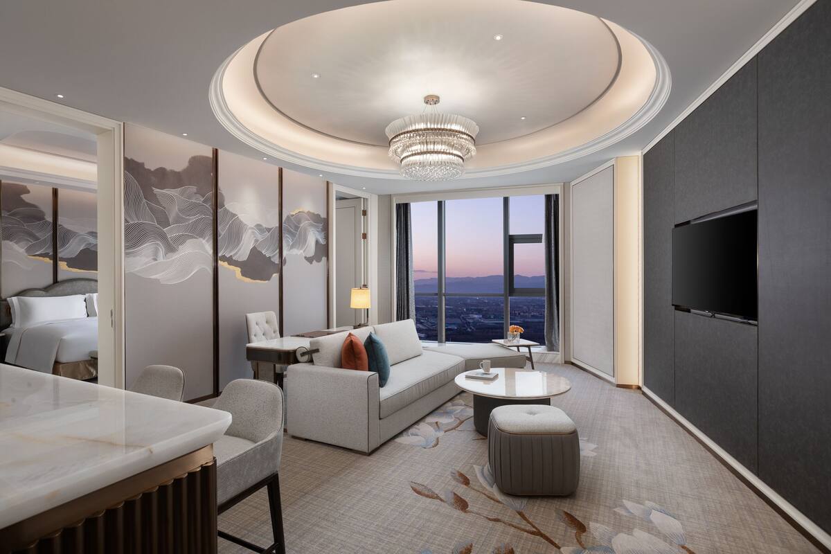 A modern hotel room with a large bed, a sitting area with a sofa and chairs, a dining table, and a large TV.