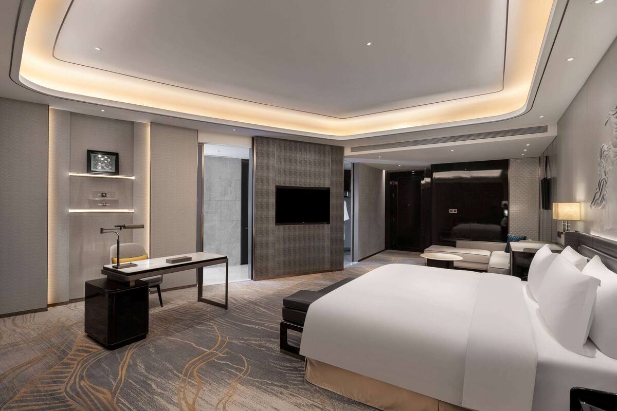 A modern hotel room with a large bed, a desk, a flat-screen TV, and a seating area with a sofa and chairs.