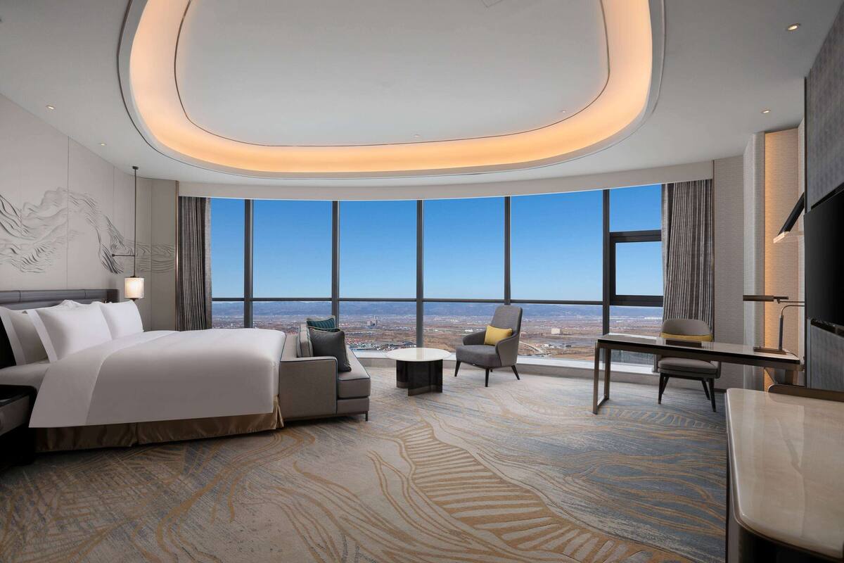 A modern hotel room with a large bed, a sofa, a desk, and a view of the city through large windows.