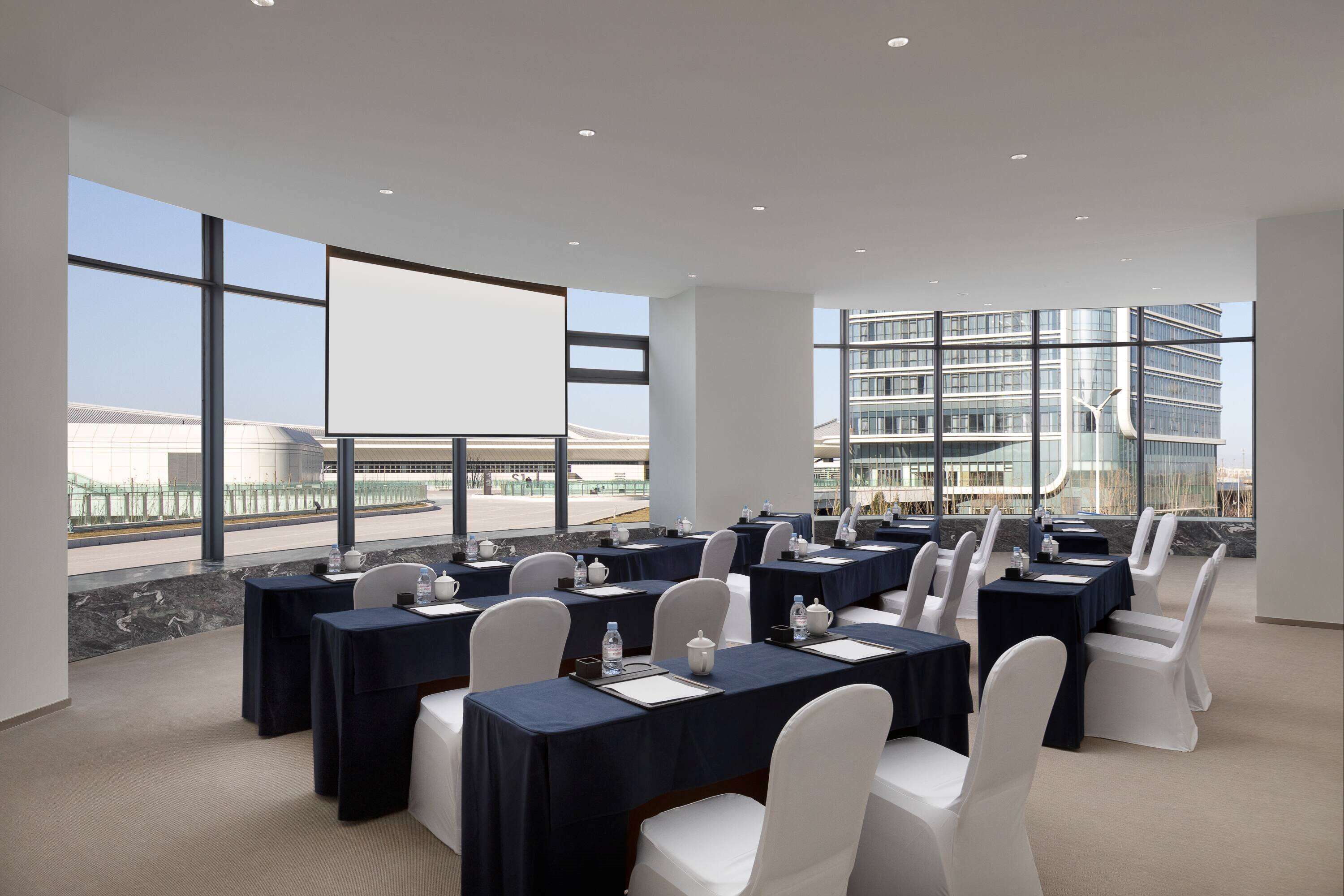 A conference room with tables set for a meeting, large windows offering views of a cityscape, and a modern building in the background.
