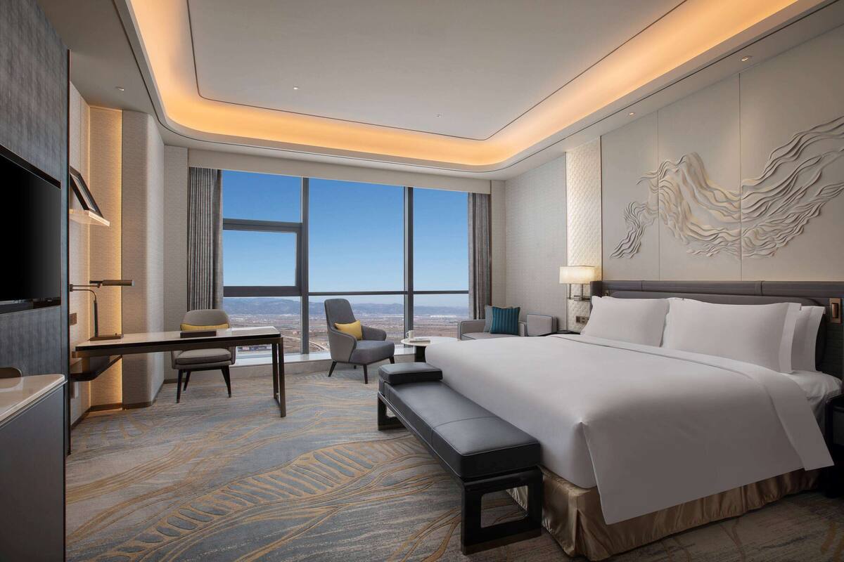A modern hotel room with a large bed, a desk, a chair, and a view of the cityscape.