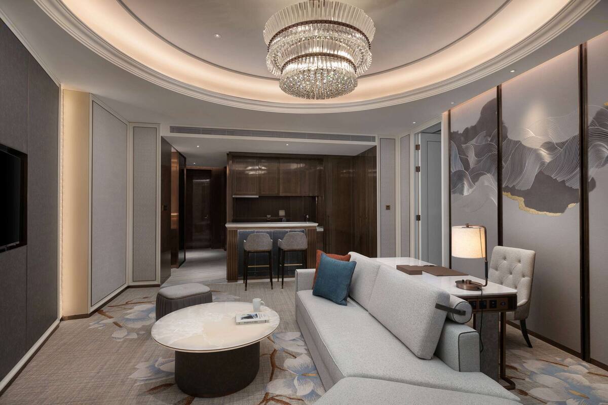 A modern hotel room with a large chandelier, a sofa, a coffee table, and a dining area.