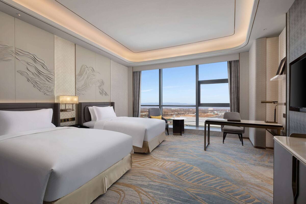 A hotel room with two beds, a desk, a chair, a TV, and large windows offering a city view.