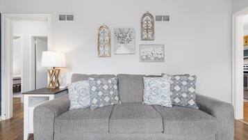 A grey sofa with patterned cushions, a small table with a lamp, and decorative wall art in a neutral-toned room.