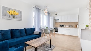 A modern living room with a blue sofa, a wooden coffee table, and a dining area with a white table and chairs. A kitchen with white cabinets and a wooden countertop is visible in the background.