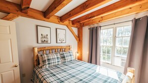 Une chambre avec un plafond en bois, un lit recouvert d’une couverture à carreaux, deux tableaux encadrés au mur et une fenêtre avec des rideaux.