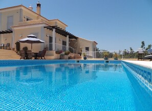 A swimming pool with a tiled edge, a covered patio area with chairs and an umbrella, and a building with a tiled roof and a balcony. - Escape & Unwind in Elegance with Large Private Pool & Amazing Views (Free Wi-Fi) (Sao Bras de Alportel, Algarve)