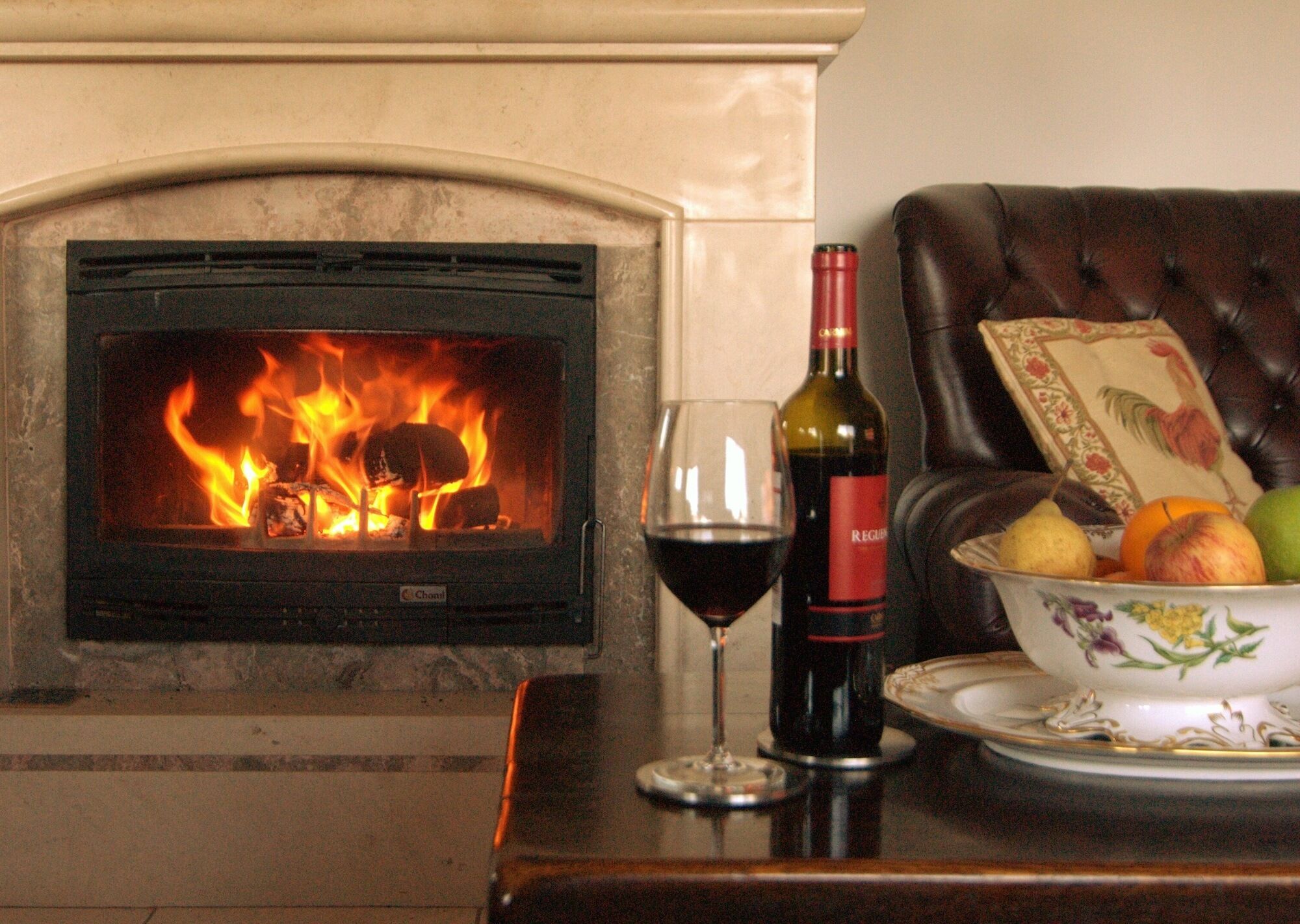 Une cheminée avec un feu crépitant, un verre de vin rouge et un panier de fruits sur une table.