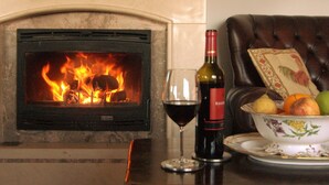 Ein Kamin mit brennendem Feuer, ein Glas Rotwein und eine Obstschale auf einem Tisch.