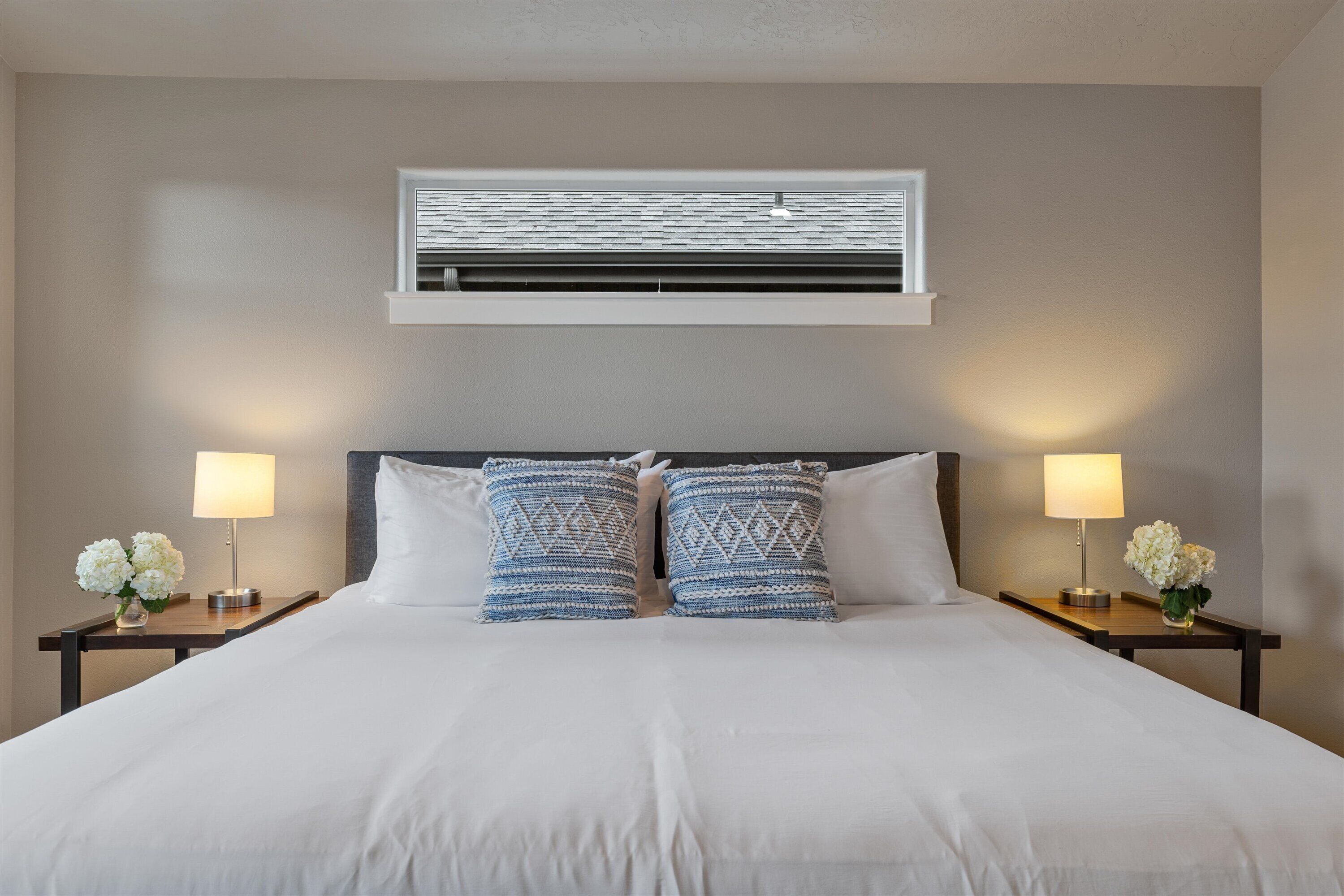 En snyggt bäddad säng med vita sängkläder, två mönstrade kuddar och två sänglampor dekorerade med vita blommor.