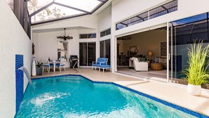 A modern indoor pool area with a glass ceiling, a seating area with chairs and a table, and a view into the living space. - Ideal Location West of 41 - Minutes to the Beaches! (Naples)