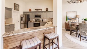A modern kitchen with marble countertops, wooden bar stools, and a dining area with a round table and chairs. - Ideal Location West of 41 - Minutes to the Beaches! (Naples)
