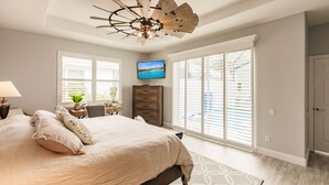 A bedroom with a large bed, a ceiling fan, a TV, and a view of the outdoors through large windows. - Ideal Location West of 41 - Minutes to the Beaches! (Naples)