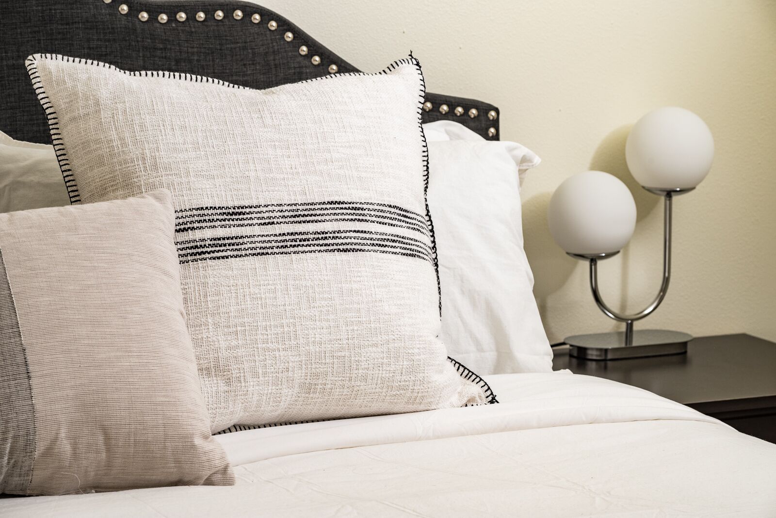 A neatly made bed with white bedding and decorative pillows, a dark headboard with metal studs, and a bedside table with two spherical lamps.