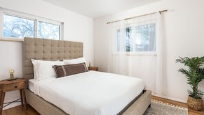 Ein Schlafzimmer mit einem großen Bett, zwei Nachttischen, einem Fenster mit durchsichtigen Vorhängen und einer Topfpflanze.