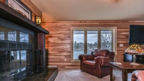 Ein gemütliches Wohnzimmer mit Kamin, Ledersofa und einem großen Fenster mit Blick auf Bäume und ein Gewässer.