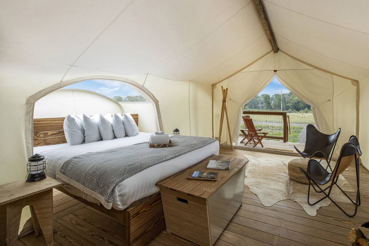 A tented room with a bed, wooden desk, chairs, and a view of the outdoors.