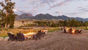 Eine Gruppe von Menschen sitzt um eine Feuerstelle in einer Outdoor-Umgebung, im Hintergrund sind Berge zu sehen.