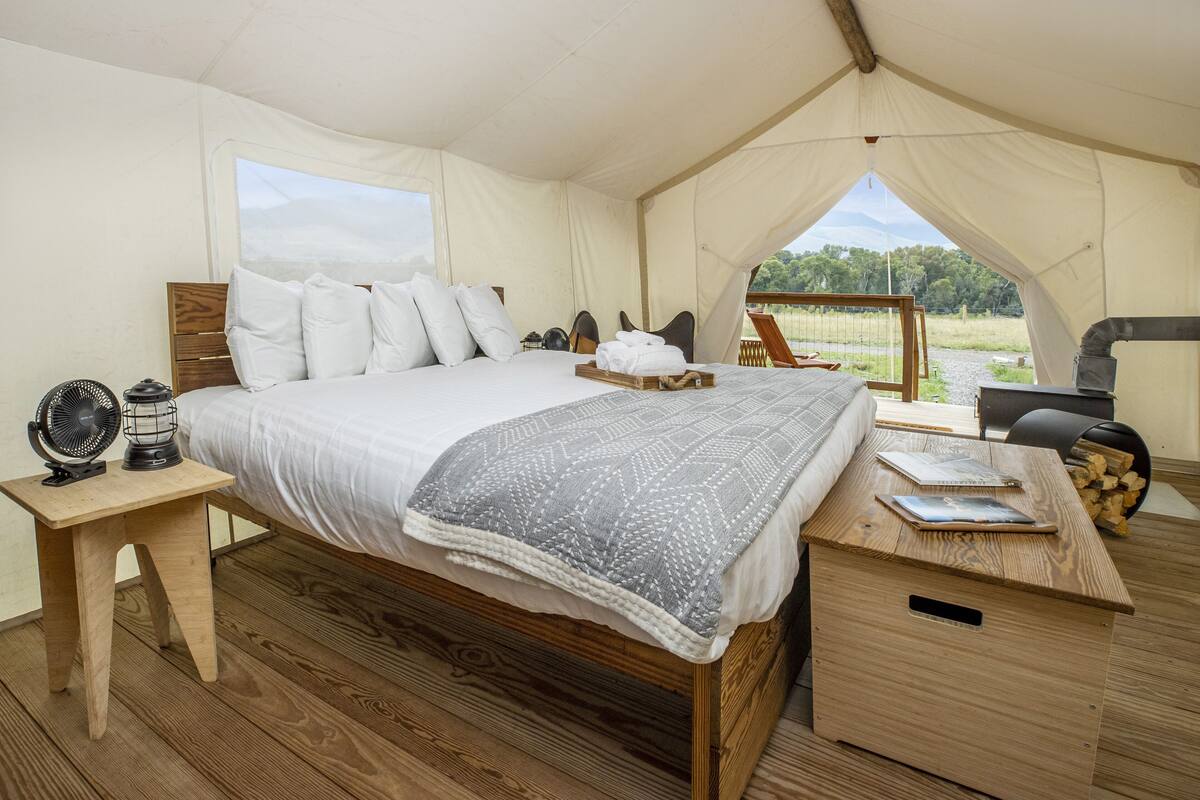 A tented room with a bed, a wooden desk, a fan, and a view of the outdoors.