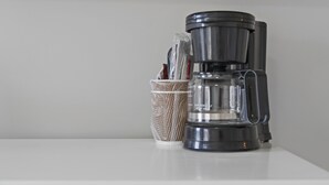 Eine Kaffeemaschine steht auf einer weißen Fläche, zusammen mit einem Korb, der eine Kapsel und einen Löffel enthält.